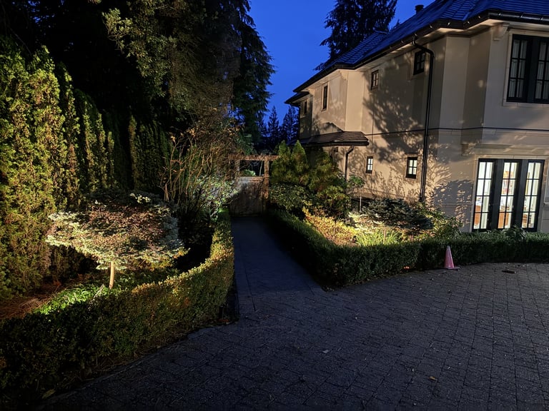 Illuminated driveway at night leading to a two-story stone house with ivy-covered hedges and lit landscaping